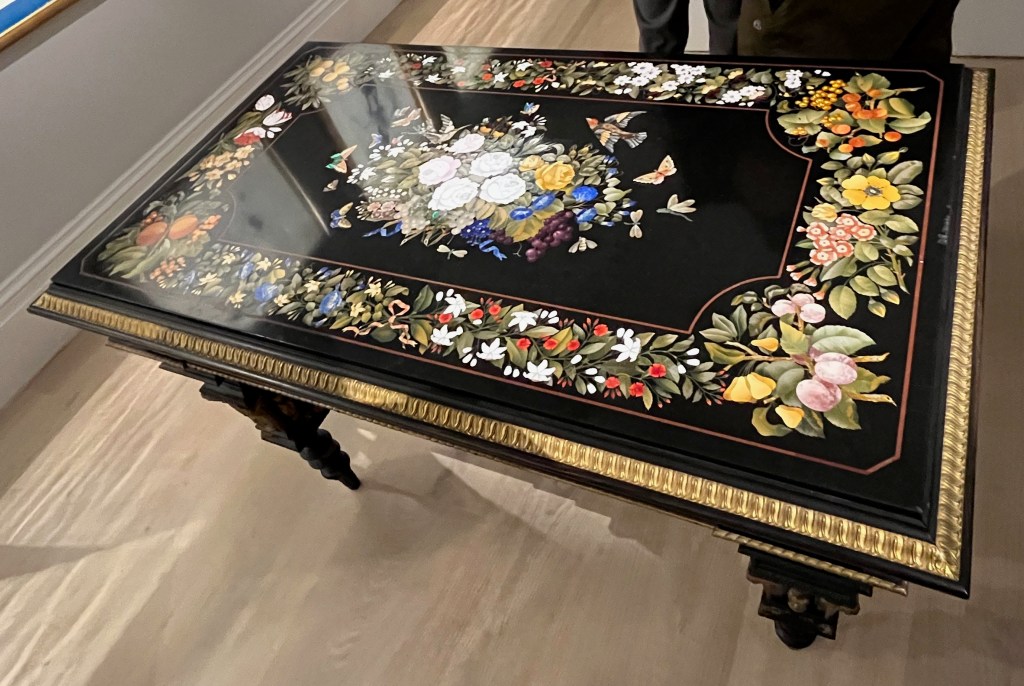 A large black table, colourfully decorated on the top. In the centre is a bouquet of ribbon-tied flowers, including roses, jasmine, pansies, bell flowers and a bunch of grapes, attracting birds, butterflies, a dragon fly and bees. Around the edge of the tabletop is a border of ribbon-tied flowers and fruit, including peaches and grapes.