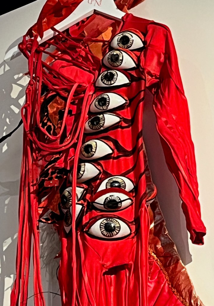 Close-up of an asymmetrical scarlet elastane catsuit, the right side and arm made of intricate red lacing, the left side with long sleeve, the stirrup legs with diagonal pleats overall in a vein-like pattern. The torso is embellished with thirteen ‘eyeballs’ of white plastic, hand-painted in black and brown, surrounded by red velvet ‘eyelids’ with hand-drawn black outlines extending up the torso to the shoulder. The back has a detachable decorative ‘spine’ made of layers of different fabrics in red, black and gold cut to resemble flames, one end with a sheaf of long plumes worn at the left shoulder, including peacock and ostrich.