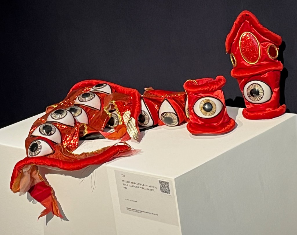 Red-coloured accessories to the outfit Freddie Mercury wore in the video for It's A Hard Life. There are two matching cuffs with Velcro fastenings. The larger right-hand cuff has a pointed end resembling a gauntlet, decorated with an ‘eyeball’ and panels of red glitter edged with gold elastic on a satin ground, edged in padded red velvet. The left-hand cuff has an ‘eyeball’ on red satin ground edged with red velvet. There are also two lower leg adornments, each with a row of six ‘eyeballs’ on a red lurex ground edged in gold braid. The reverse is cut to resemble a vertical row of four flames, with ‘silver’ tips of fabric resembling fish scales concealing the Velcro fastening. They are trimmed at the top and bottom with padded red velvet.