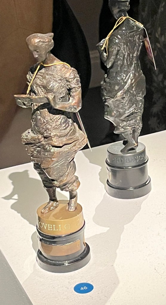 Two Ivor Novello awards, one viewed from the front, the other from the back, each consisting of a bronze statuette of Greek muse Euterpe on a circular black wooden plinth.