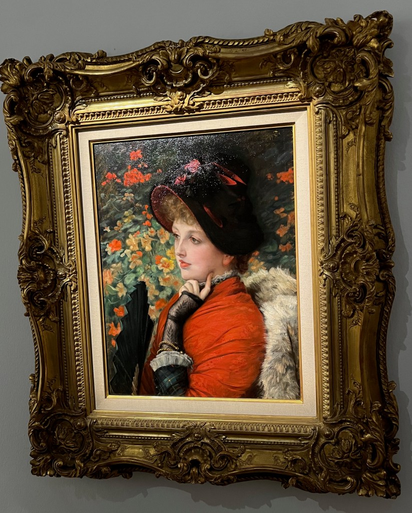 A painting called Type of Beauty, Portrait of Mrs. Kathleen Newton, by James-Jacques-Joseph Tissot. It is a half-length portrait of a woman posing and looking thoughtful, resting a finger on her lower left cheek, while wearing a black hat and a red top, in front of a flowery background of nasturtiums and geraniums.
