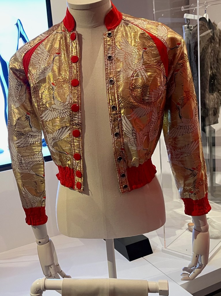 A lavish jacket trimmed in scarlet, the sparkling gilt fabric woven with an elaborate pattern of gold and white red-capped flying cranes, which are highly auspicious Japanese symbols. The jacket also has a scarlet crêpe collar, upper sleeve panels, and elasticated cuffs and hem. It fastens with scarlet buttons and is finished with scarlet stitching to the seams.