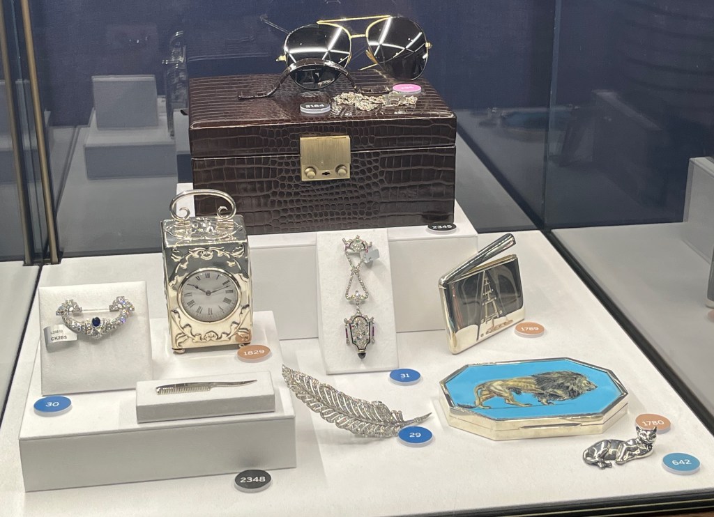 A glass case containing a variety of items including a jewellery case, aviator sunglasses, a pendant with a letter F on it, A sapphire and diamond brooch, a silver Victorian clock decorated with small cherubs and foliage, a silver moustache comb, a ruby, sapphire, emerald, onyx and diamond fob watch, a diamond brooch in the shape of a feather, a silver card case with a triangle design, and an octagonal-shaped silver and enamel box with an image of a lion against a blue background.