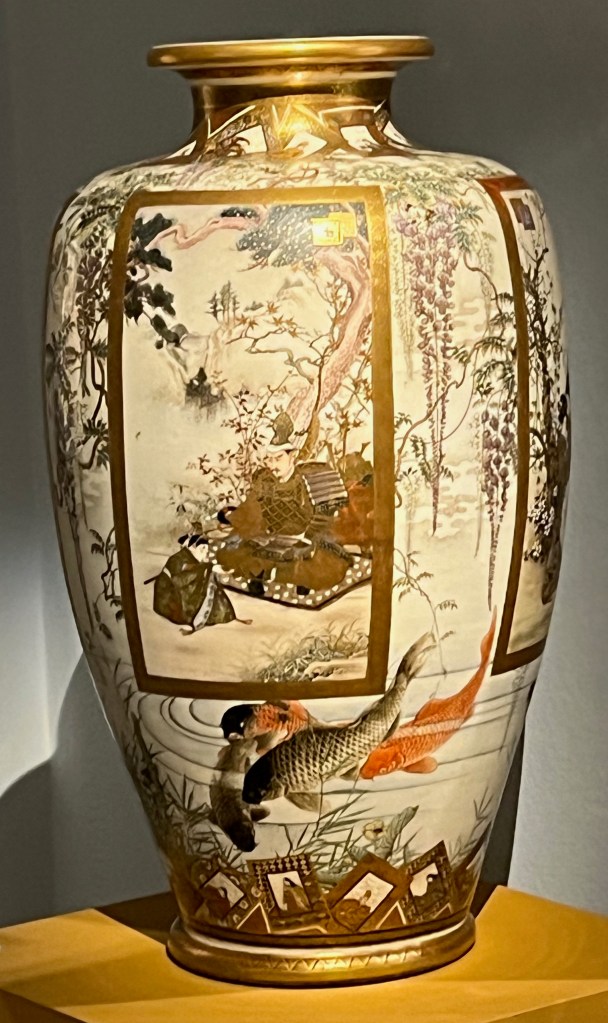 A large Japanese vase with ornately designed panels all the way around it. The one facing us depicts a samurai and a young retainer seated beneath pine and maple trees. A group of 4 fish are swimming by beneath the panel.