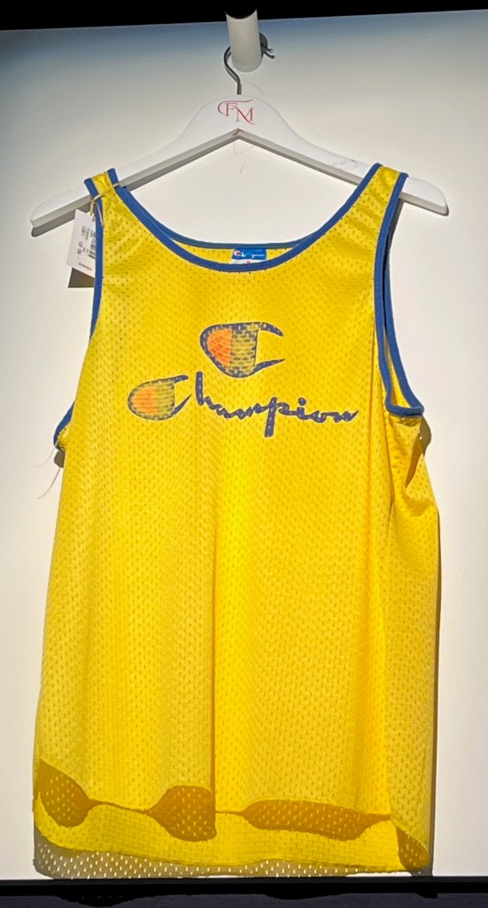 An athletics vest of yellow mesh edged in royal blue cotton, the front decorated with brand lettering and logo ‘Champion’ in royal blue with a touch of red, though the blue on the letter C of the logo is a bit faded.