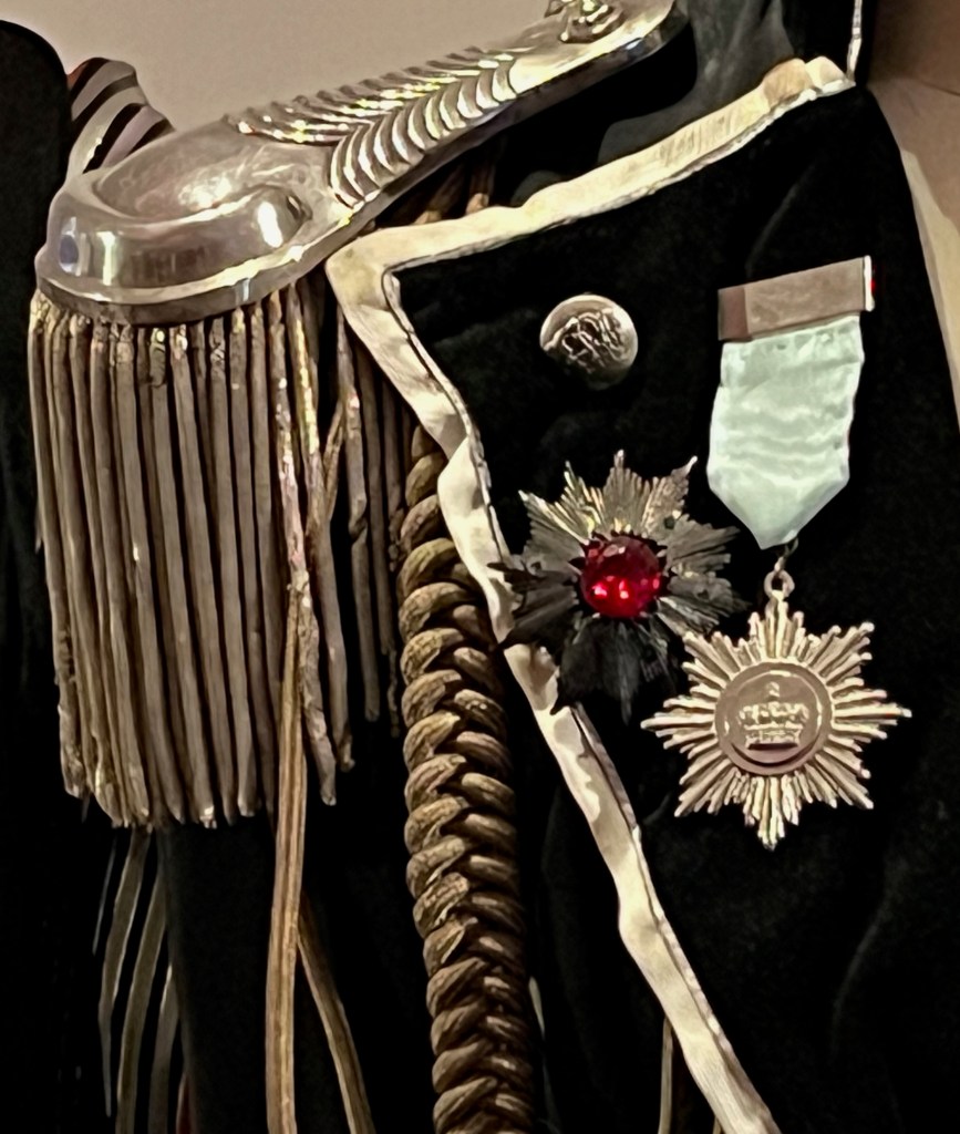 Close-up of a highly ornate short fitted jacket in a ceremonial military style. It shows a large silver shoulder epaulettea with a rose button and silver coil fringing. An on the chest next to it are two white metal imitation star medals, one centred with a red jewel-like cabochon, the other with an embossed crown motif.