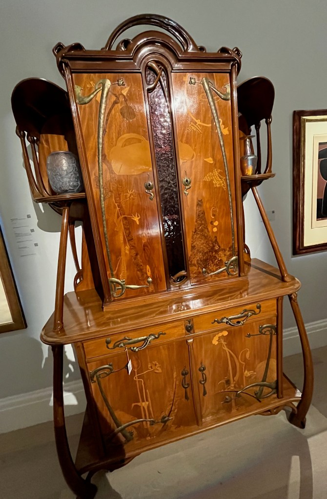 A large wooden sideboard with a 2 door cabinet at the bottom, and a much taller but slightly less wider 2-door cupboard on the top. 2 small shelves also stick out from either side of the tall cupboard, holding small silver vases.