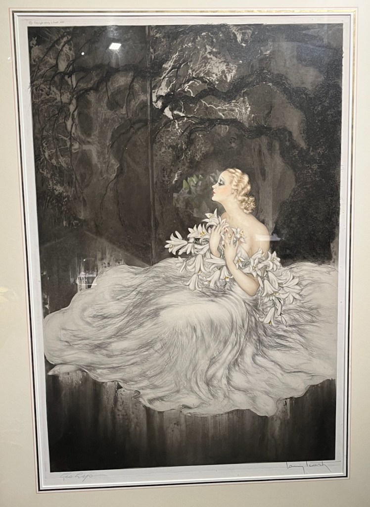 Lilies, a print by Louis Icart that shows a lady in a long flowing white dress, sat on the ground so that the skirt of the dress spreads out widely around her. She is looking thoughtful as she holds a bunch of white lilies up to her chest.