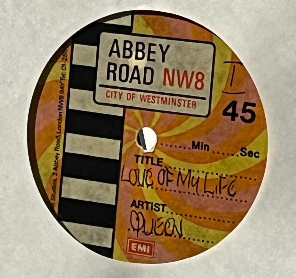 A 7-inch acetate record for Love Of My Life by Queen. At the top centre of the disc is the Abbey Road logo, which is an image of a City of Westminster street sign for Abbey Road, NW8. Down the left side of the disc is a series of black and white stripes to resemble the famous Abbey Road zebra crossing. The song title and artist are handwritten on the relevant lines in the bottom right section. The background is a swirling pattern of yellow, orange and pink lines.