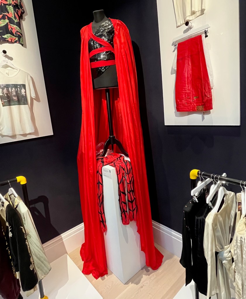 The elaborate red and black outfit worn by Freddie Mercury in his video for Made In Heaven. It includes a black asymmetrical vinyl bodice with one left sleeve ending in a point and fastening at the wrist with vinyl straps, the front covered with diagonal lines of black stitching with long straps to tie around the waist. Draped over this is a voluminous red silk cape, designed to be worn diagonally across the back, gathered at the neck and trimmed with red jersey fabric, with long red straps wrapping under the arms and around the torso to fasten.