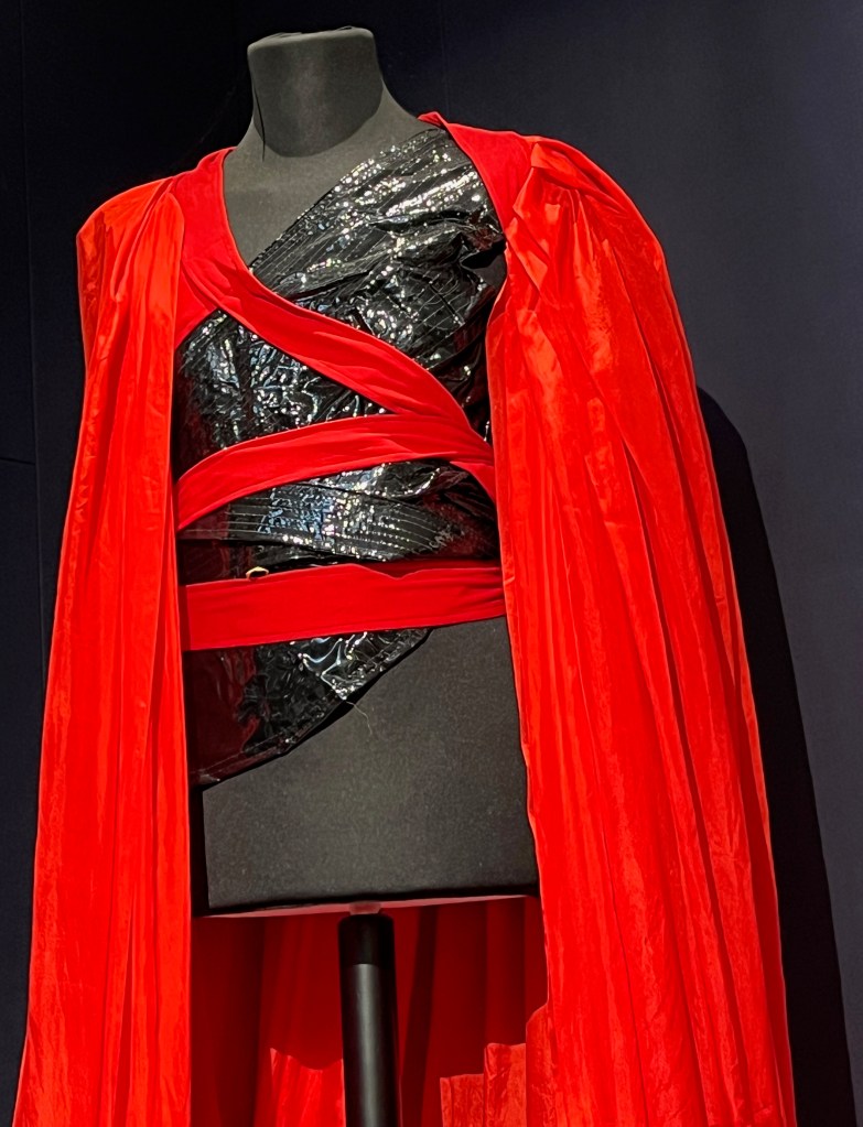 Close-up of the elaborate red and black outfit worn by Freddie Mercury in his video for Made In Heaven. It includes a black asymmetrical vinyl bodice with one left sleeve ending in a point and fastening at the wrist with vinyl straps, the front covered with diagonal lines of black stitching with long straps to tie around the waist. Draped over this is a voluminous red silk cape, designed to be worn diagonally across the back, gathered at the neck and trimmed with red jersey fabric, with long red straps wrapping under the arms and around the torso to fasten.