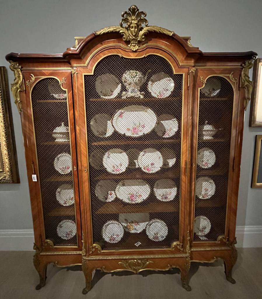 A large, ornate, French kingwood bookcase. It has a large central door and 2 much narrower side doors, with all doors having a grille effect, slightly obscuring the items inside. White plates of various sizes, with small floral designs on them, having been placed on the 5 shelves inside. The pediment on the top of the bookcase has a golden female mask with 5 curling feather-like structures over her head.