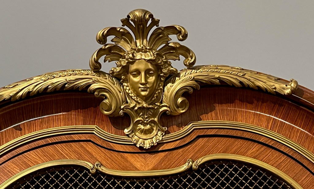 Golden pediment on top of a wooden bookcase, in the form of a golden female mask with 5 curling feather-like structures over her head.