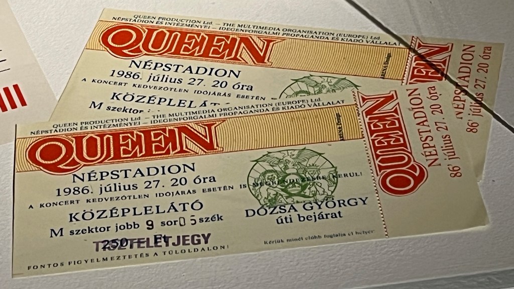 Two tickets to see Queen at Népstadion, Budapest, Hungry on 27 July 1986. The name Queen is in large red letters with white edging along the top and down the side of the ticket. The ticket also includes an image of the band's crest in green ink.