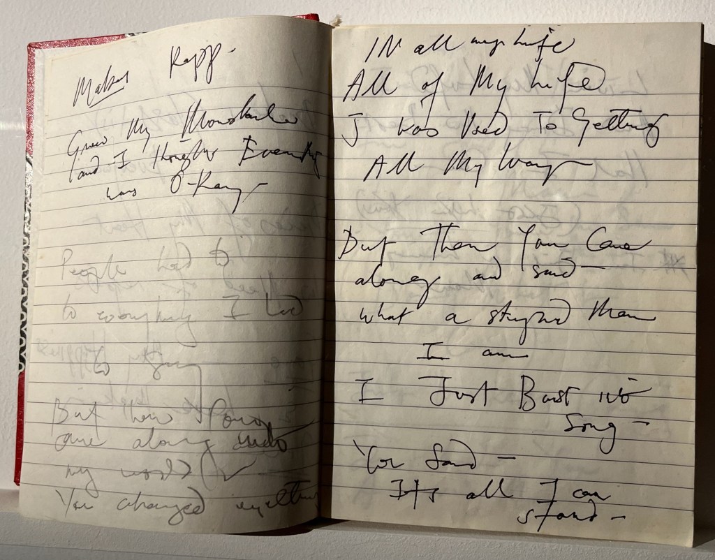 A double-page spread from a notebook with handwriting by Freddie Mercury. The left page has the draft lyrics Grew my Moustache and I thought Everything was O-Kay. On the right hand page, the lyrics include In all of my life, all my life, I was used to getting all my way. But then you came along and said what a stupid man I am. I burst into song. You said it's all I can stand.