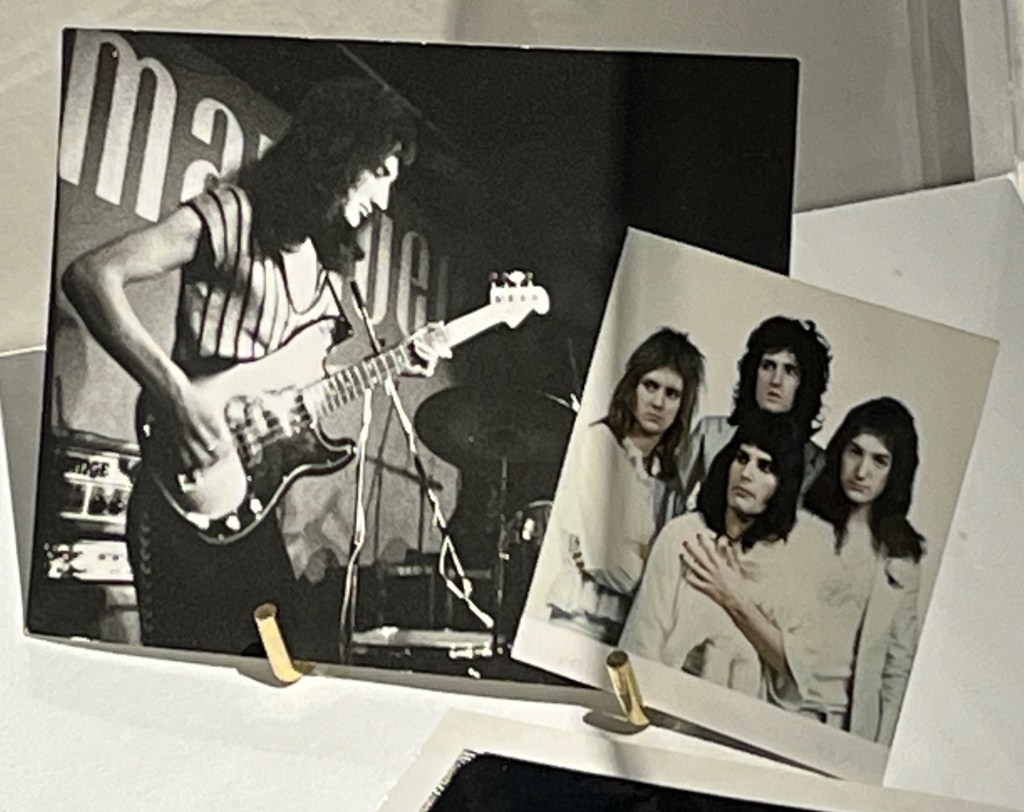 Two black and white polaroid photos, one showing Queen bass guitarist John Deacon playing on a stage, the other showing the 4 members of Queen posing together.
