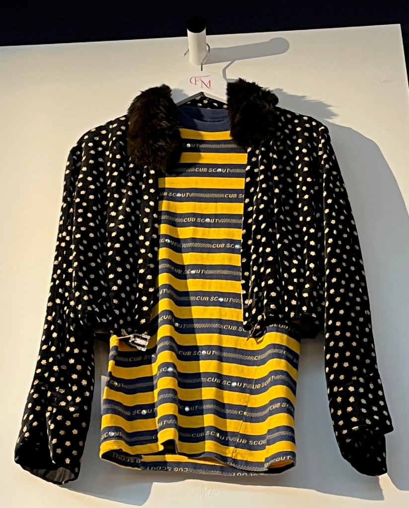 A cropped black velvet vintage jacket printed with a pattern of gold flowerheads, with a fur collar, flared sleeves, and the open front with art deco diamante clip fastening at the waist. The material has three pleats either side of the front fastening, lined in cream silk, the cuffs additionally lined with black silk. This is hung over a blue and yellow striped T-shirt, the blue stripes woven with the words ‘CUB SCOUT’ in yellow and studded with diamante.