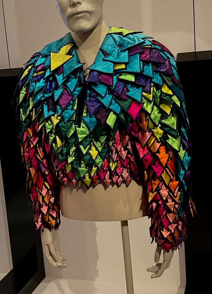 A colourful jacket, cut in short matador style. It has a purple satin ground covered with numerous padded satin arrows in a myriad of bright colours edged in black, the arrows stitched individually in a dense cascade pattern descending in size from the shoulder to the hem and giving an overall effect of movement, similar to the feathers of an exotic bird, lined in black satin and fastening at the front with Velcro.