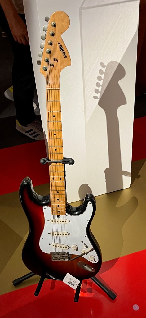 A Satellite model of guitar, with maple neck and a 21-fret fingerboard with dot markers. The body of the guitar is dark brown around the edges of the central white design.