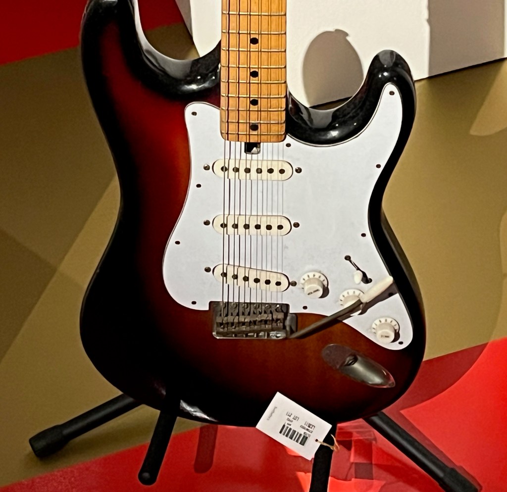 Close-up of the body of a Satellite model of guitar, which is dark brown around the edge of the white central section, with a maple neck sticking up from it.