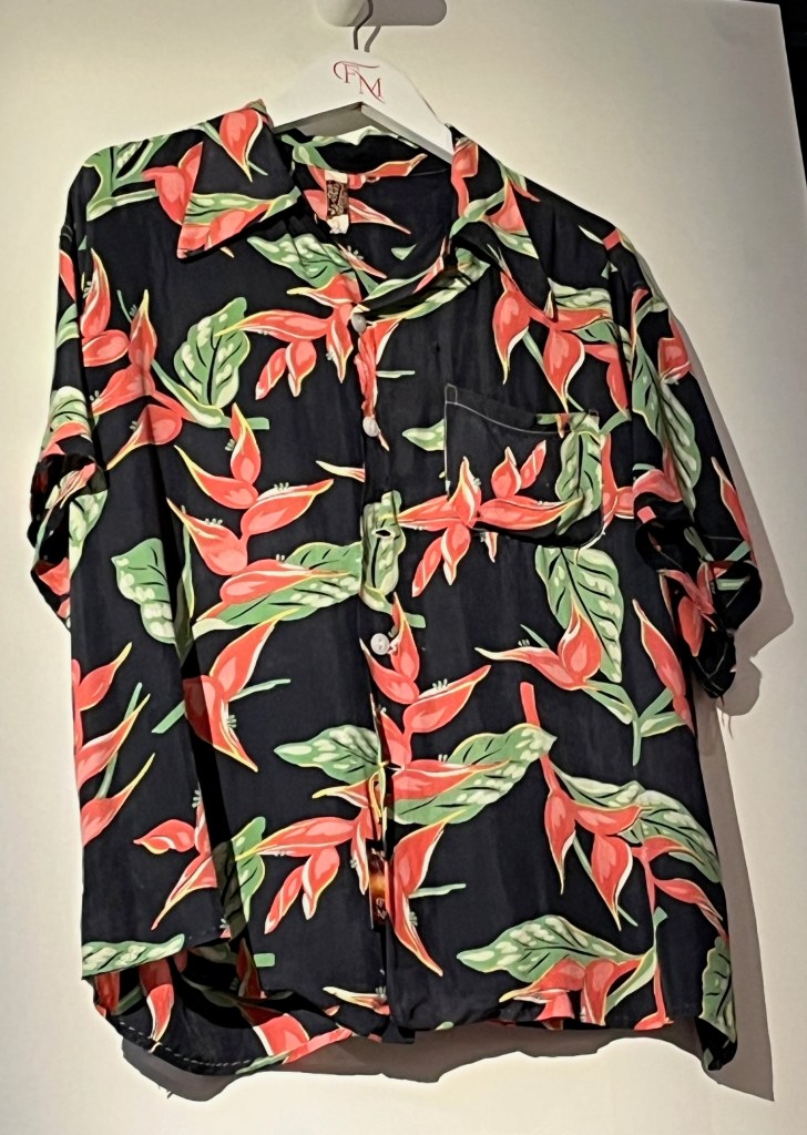 A short-sleeve Hawaiian-style shirt with pointed collar. The black background is printed with a pattern of bird of paradise flowers in bright pink and green. There is also a left breast pocket, and the shirt is fastened with white plastic buttons.