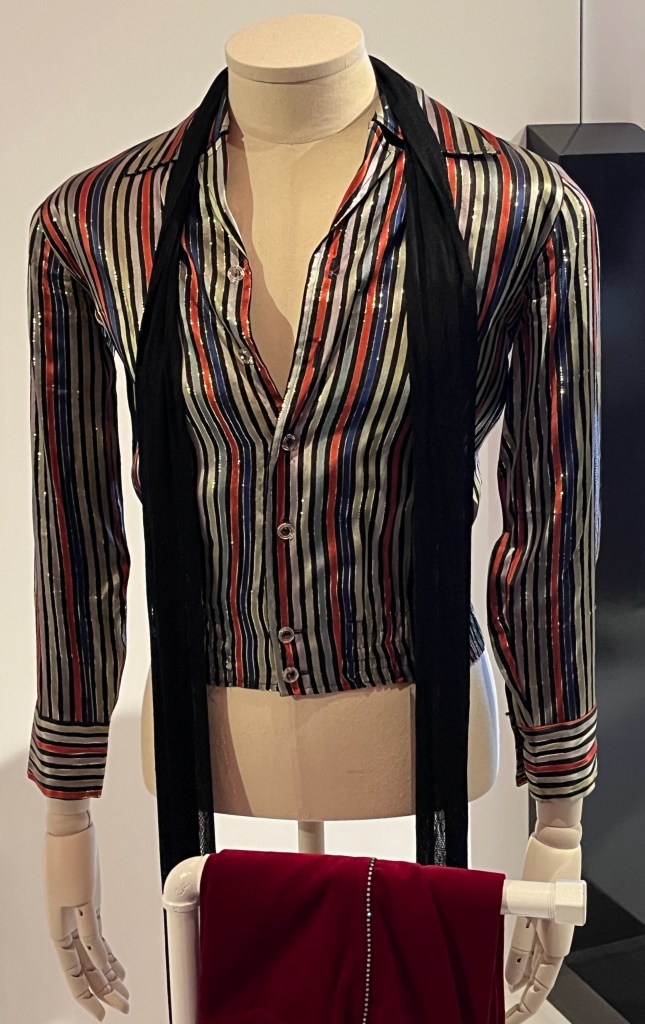 A jacket made of black, red, blue and olive striped lurex with pointed collar and elasticated hem, the front and cuffs fastening with circular clear plastic buttons.