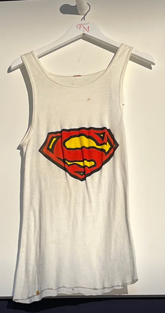 A white cotton ribbed vest with the Superman logo hand-stencilled in red, yellow and black ink.