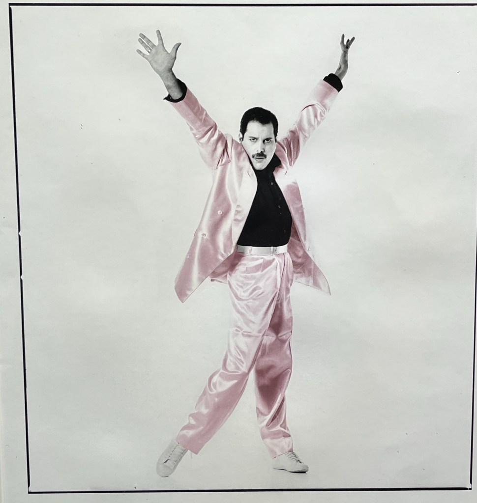 A black and white promotional photo for Freddie Mercury's song The Great Pretender. Against a plain white background, it shows Freddie in a shiny jacket and trousers, the jacket open to reveal a black top beneath. He's posing flamboyantly with his arms above his head in a V-like shape.