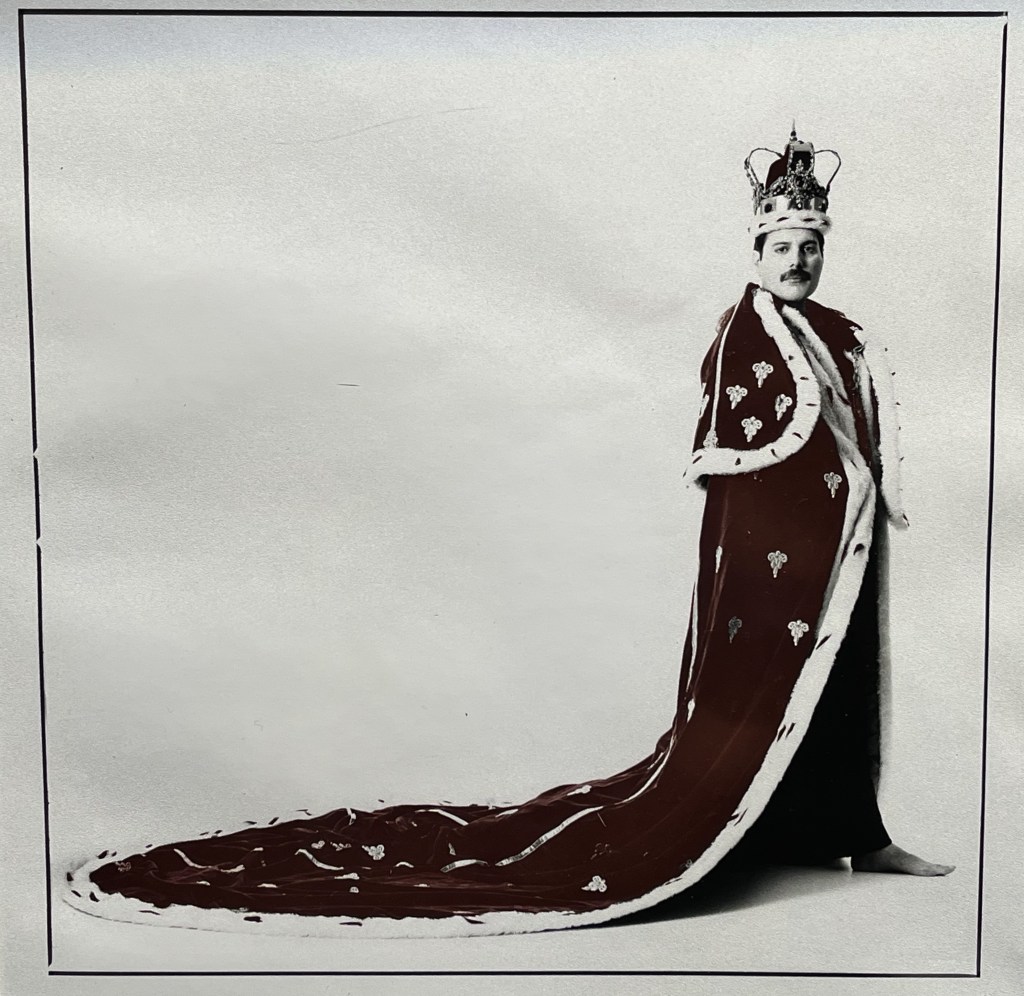 A black and white promotional photo for Freddie Mercury's song The Great Pretender. Against a plain white background, it shows a side view of Freddie standing in his ornate royal robe and crown, the large train of the robe spread out behind him.