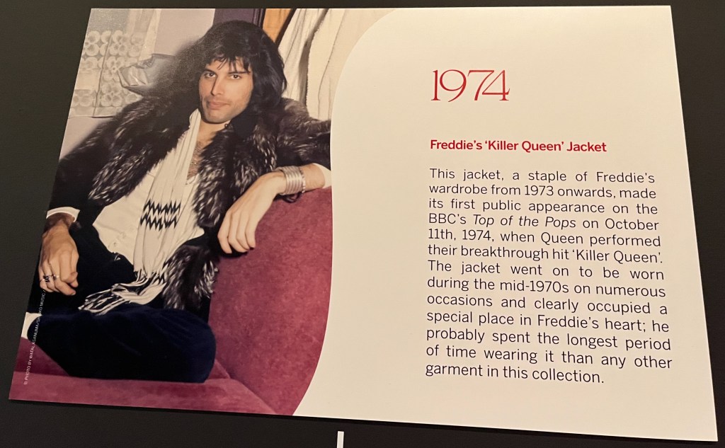 A white board on the Freddie Mercury timeline in Sotheby's. Alongside a photo of Freddie Mercury sitting casually while wearing a fur jacket and a white scarf is text that reads 1974 - Freddie's 'Killer Queen' Jacket. This jacket, a staple of Freddie's wardrobe from 1973 onwards, made its first public appearance on the BBC's Top of the Pops on October 11th, 1974, when Queen performed their breakthrough hit 'Killer Queen'. The jacket went on to be worn during the mid-1970s on numerous occasions and clearly occupied a special place in Freddie's heart; he probably spent the longest period of time wearing it than any other garment in this collection.