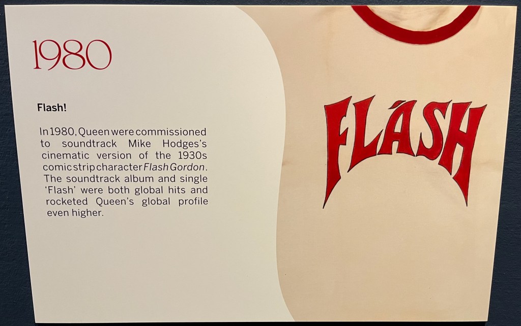 A white board on the Freddie Mercury timeline in Sotheby's. Alongside a close-up picture of the large red Flash logo from Flash Gordon on a white t-shirt is text that reads 1980 - Flash. In 1980, Queen were commissioned to soundtrack Mike Hodges's cinematic version of the 1930s comic strip character Flash Gordon. The soundtrack album and single 'Flash' were both global hits and rocketed Queen's global profile even higher.