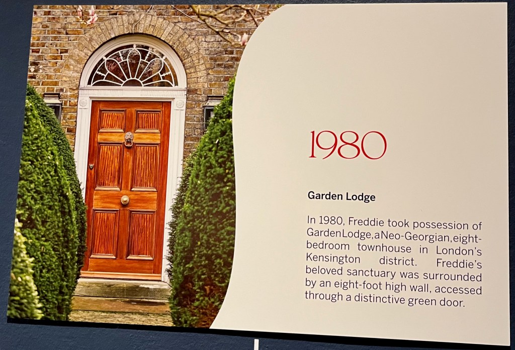 A white board on the Freddie Mercury timeline in Sotheby's. Alongside a picture of the wooden front door of Garden Lodge, with bushes each side of the narrow pathway, is text that reads 1980 - Garden Lodge. In 1980, Freddie took possession of Garden Lodge, a Neo-Georgian, eight-bedroom townhouse in London's Kensington district. Freddie's beloved sanctuary was surrounded by an eight-foot high wall, accessed through a distinctive green door.