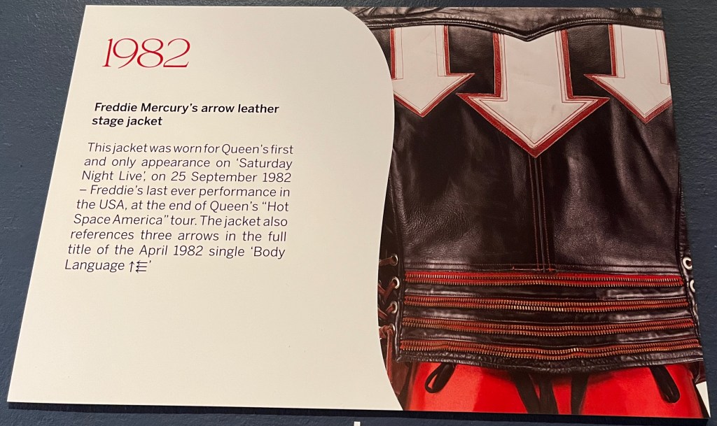 A white board on the Freddie Mercury timeline in Sotheby's. Alongside a close-up picture of 3 large white arrows pointing down on a black leather jacket is text that reads 1982 - Freddie Mercury's arrow leather stage jacket. This jacket was worn for Queen's first and only appearance on 'Saturday Night Live', on 25 September 1982 - Freddie's last ever performance in the USA, at the end of Queen's "Hot Space America" tour. The jacket also references three arrows in the full title of the April 1982 single 'Body 'Language', the title finishing with an up arrow followed by a set of 3 left-facing arrows one above the other.