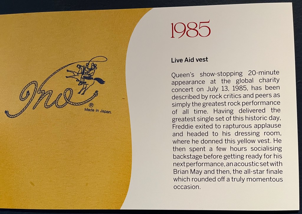 A white board on the Freddie Mercury timeline in Sotheby's. Alongside a close-up of a black drawing of a cowboy on a horse swinging a lasso on a yellow t-shirt is text that reads 1985 - Live Aid vest. Queen's show-stopping 20-minute appearance at the global charity concert on July 13, 1985, has been described by rock critics and peers as simply the greatest rock performance of all time. Having delivered the greatest single set of this historic day, Freddie exited to rapturous applause and headed to his dressing room, where he donned this yellow vest. He then spent a few hours socialising backstage before getting ready for his next performance, an acoustic set with Brian May and then, the all-star finale which rounded off a truly momentous occasion.