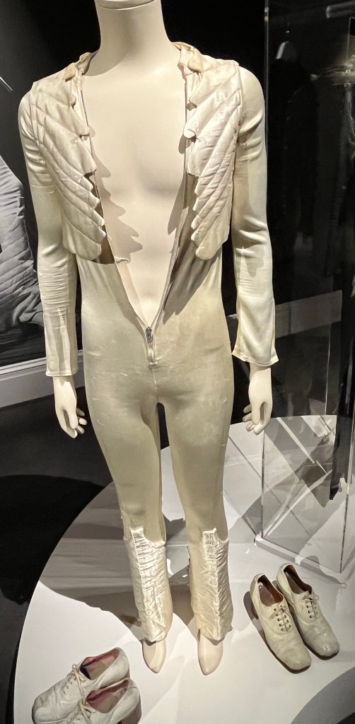 White catsuit with deep-cut zip-fastening front from waist to curved stand collar. The long narrow sleeves are zipped at the cuffs and embellished with four quilted panels terminating in points to resemble bird wings. The slightly flared trouser legs are similarly applied with quilted bird-wing adornments, and these wrap-around trouser leg panels graduate in width from their apex below the knee to their widest point at the foot. Each trouser leg also has a stirrup of ribbed webbing at the base,