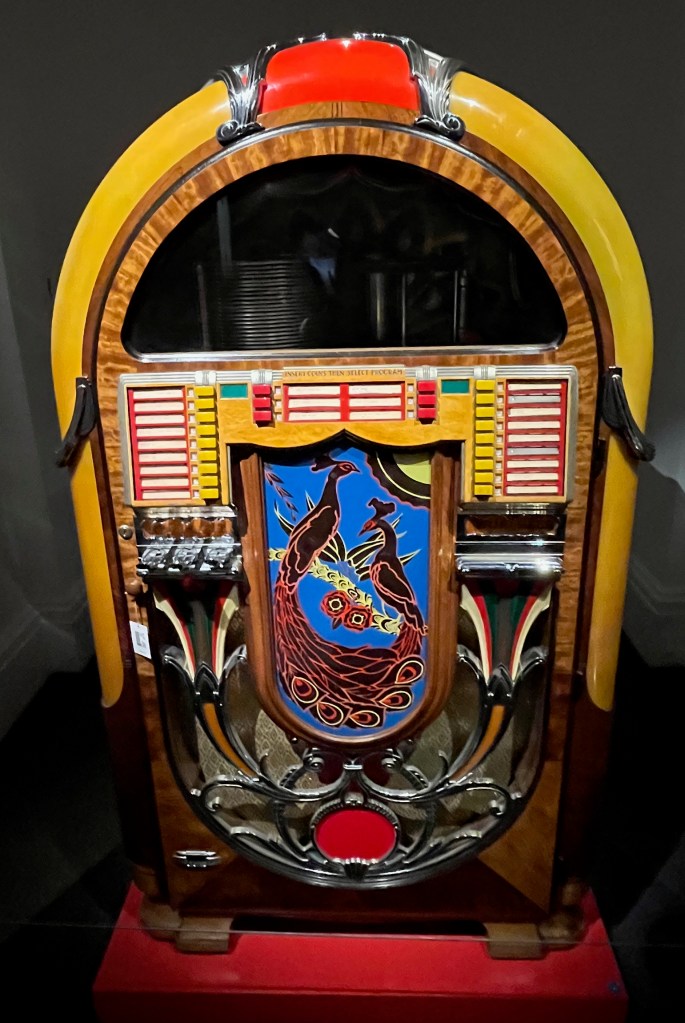 A colourful Wurlitzer jukebox, with buttons for 24 records, and a curved panel on the front showing stylised artwork of 2 black peacocks with red edging to their plumage, perched in front of a blue background.