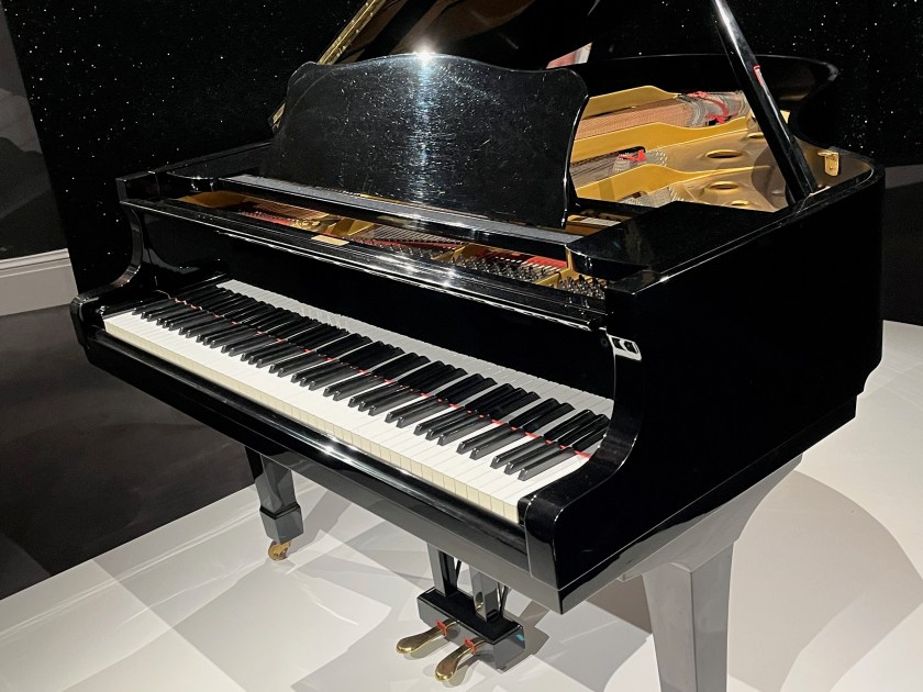 Freddie Mercury's black baby grand Yamaha piano, viewed from an angle to the right so the strings are visible beneath the lifted lid at the back.