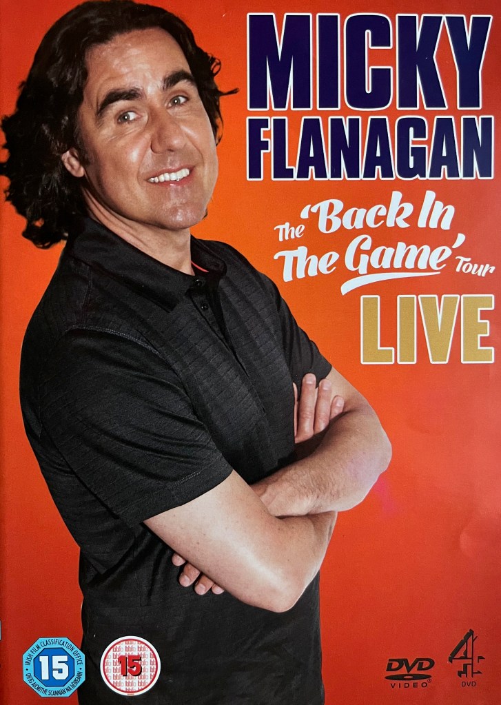 Front cover of the DVD for Micky Flanagan, Back In The Game Live, showing a picture of the comedian wearing a black shirt, standing with his arms folded across his chest and a smile on his face.