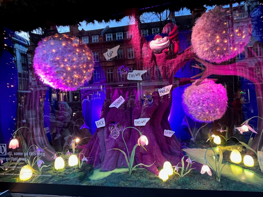 Selfridges window display representing Alice In Wonderland, showing a colourful forest scene with the grinning Cheshire cat looking out from high up in one of the trees. A selection of white signs dotted around the scene point in different directions, with words and phrases like up, down, this way, yonder and back.