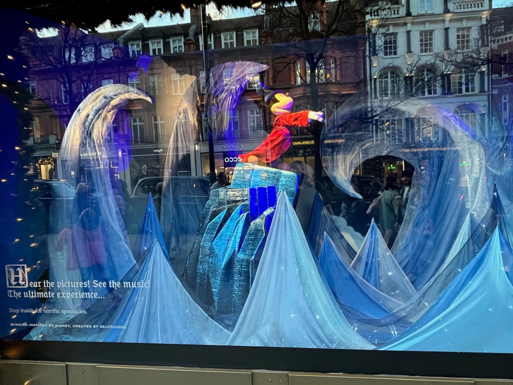 Selfridges window display representing Fantasia, with Mickey Mouse standing on a tall rock wearing a red cloak and a black wizard's hat, his hands outstretched as he controls the large curving waves of water that are towering over him.