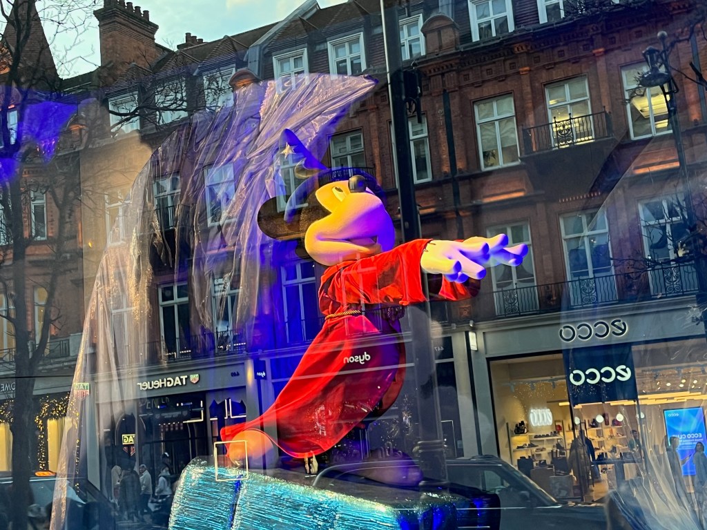 Close-up of Mickey Mouse standing on a tall rock wearing a red cloak and a black wizard's hat, his hands outstretched as he casts a spell, in a Selfridges window display representing Fantasia.