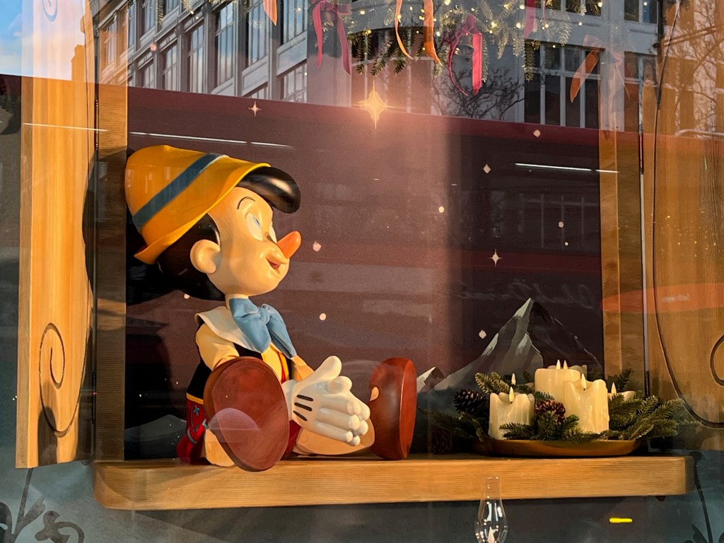 Close-up of the Pinocchio puppet on a shelf, in a Selfridges window display representing the Disney film. He's wearing a yellow hat and top, a blue bow tie, white gloves and brown shoes.