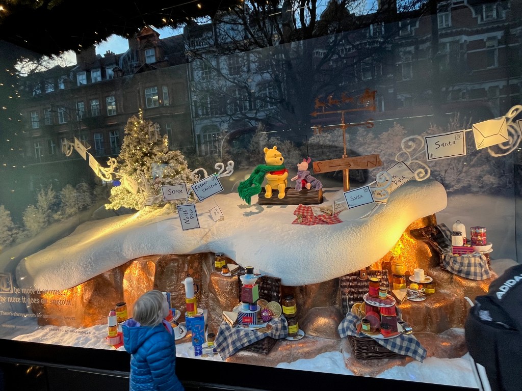Selfridges window display representing Winnie The Pooh, showing the character sitting on a log with piglet in the middle of a snowy landscape with a decorated Christmas tree nearby. A length of string running across the scene has envelopes attached to it addressed to Santa, Father Christmas and the North Pole. below the landscape, as if buried underground, is a huge amount of food.