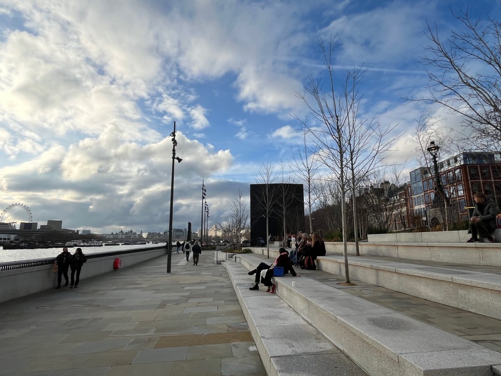The Bazalgette Embankment by the River Thames. On the left, people walk along a path by a low wall, with a couple stopping to admire the view of the river over it. On the right, some people are sitting on large concrete steps, which have a few thin trees without leaves sticking up from them. At the far end of the steps is the back of a large black monolith structure.