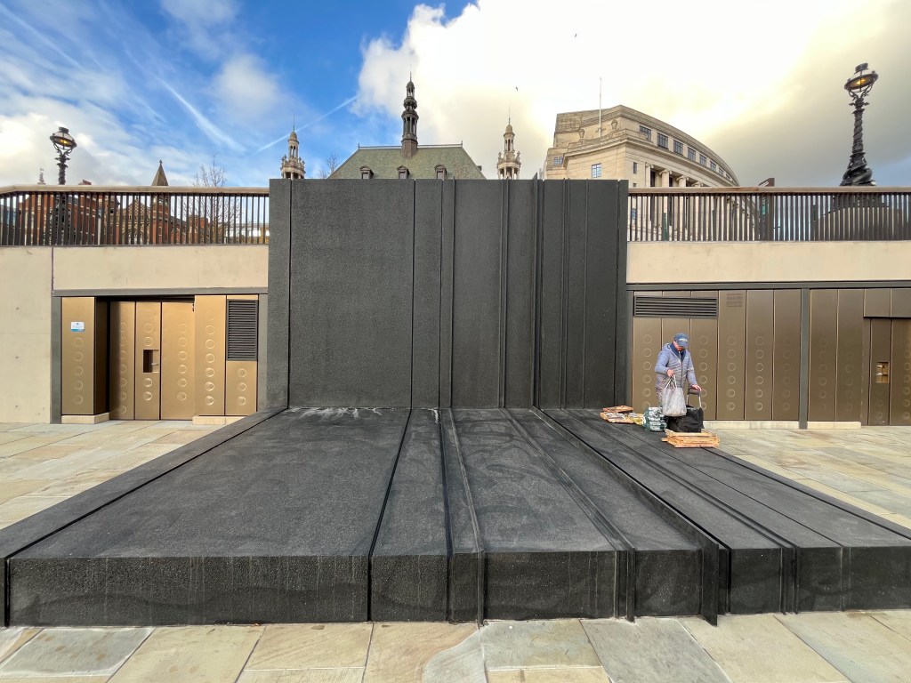 A very large and wide black monolith structure, which has a section stretching outwards on the ground as well as up the wall and railings at the back. At random intervals across the structure are ridges or raised lines that span its full length.