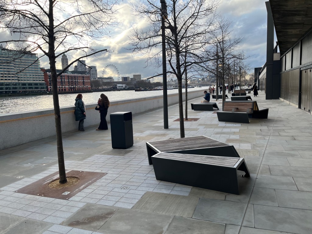 Bazalgette Embankment, with people walking along the pathway or stopping to look at the River Thames over the low wall. There are also some benches for people to sit on, interspersed with some thin, bare trees.