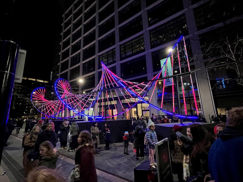 Blueprint, an artwork lit up in purple and pink colours that looks like a double helix strand of DNA, with 2 spiralling lines linked by straight lines.