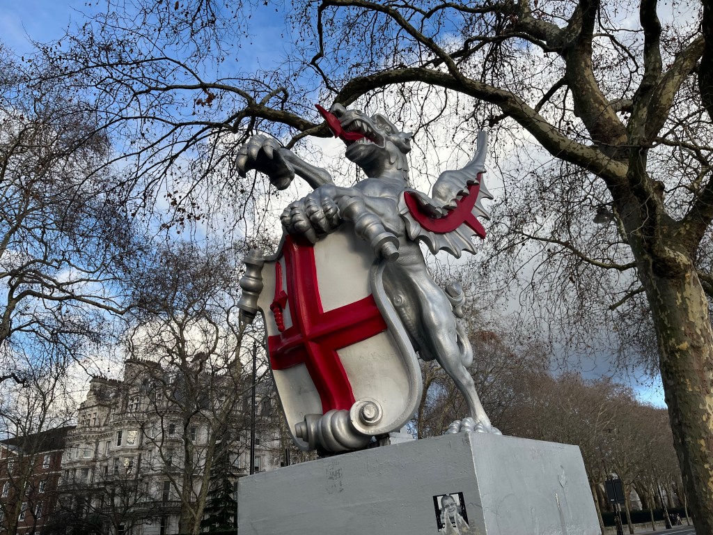 A sculpture of a dragon on a plinth, serving as a boundary marker for the City of London. The dragon is painted silver, with a red tongue and some red detailing under its wing. It's standing on its left rear leg, while the right rear leg is lifted forward to support the back of a shield. Its left front leg is holding the top of the shield to keep it upright, while its right front leg is raised in the air. The shield has the flag of St George, a red cross on a white background, plus a red sword in the top left quarter of the flag.