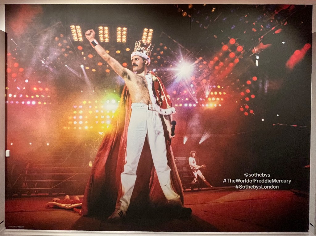 Freddie Mercury posing proudly on stage, pumping his right fist into the air. Bare-chested Freddie has a royal red robe with gold decoration and white fur trim draped over his shoulders, and an ornate gold, gem-studded crown with white fur trim on his head. He's also wearing white trousers. In the background, beneath the bright colourful lights that illuminate the stage, Brian May is standing with legs astride as he plays the guitar.