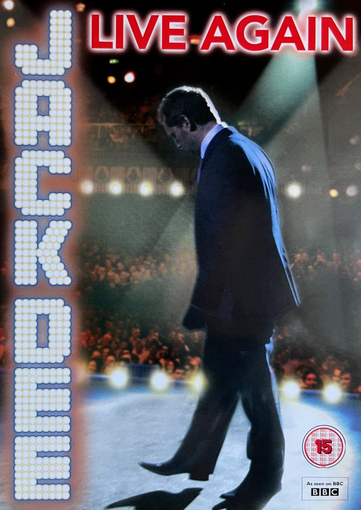 Front cover of the DVD for Jack Dee Live Again. It shows a side view of Jack, head slightly bowed, as he walks across the stage, lit by a spotlight from high above. The scene is viewed from the back of the stage so the audience are in the background. His name is down the left side of the cover, each letter made up of lots of little spotlights, like the ones pointing at him along the edge of the stage and across the upper circle in the auditorium. The title live again is in red letters across top above Jack.